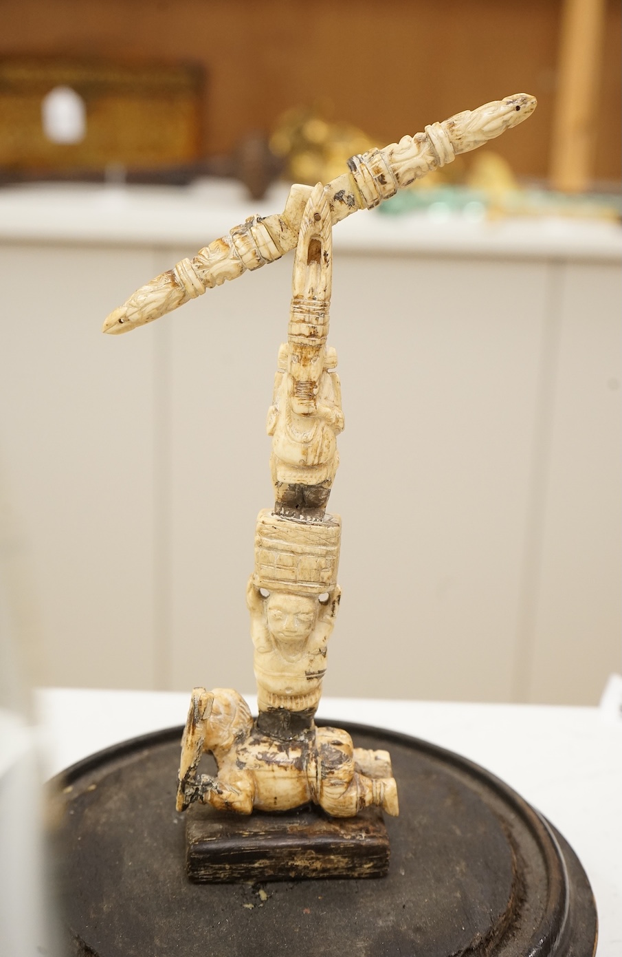 A 19th century Indian bone sculptural devotional object, carved as a totem with Ganesh and other deities, pivoting beam, 18cm high, glass dome with ebonised base